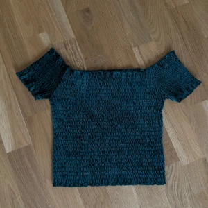 Mörkgrön smockad offshoulder topp - Trendig mörkgrön topp med smockad struktur och offshoulder-design. Kortärmad och croppad modell som sitter tight och ger en snygg siluett. Perfekt för dig som vill ha en cool och bekväm look.