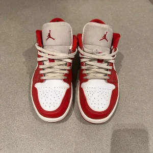 Nike Air Jordan 1 Low röd/vit - Snygga Nike Air Jordan 1 Low sneakers i rött och vitt skinn med klassisk röd swoosh och Jumpman-logga på plösen. Lågt skaft, perforerad tå och vita snören. Perfekta för dig som vill sticka ut med en ikonisk streetstyle.