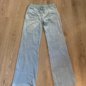 Ljusblå velourbyxor Juicy Couture - Säljer ett par ljusblå mjukisbyxor från Juicy Couture i mjuk velour. Byxorna har raka ben, snörning i midjan och stora fickor fram och bak med snygga detaljer i form av knappar och broderad logga på ena fickan.