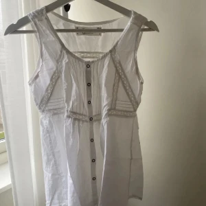 Vit blus från RoseBud med spetsdetaljer - Supersöt vit blus från RoseBud i tunn bomull med spetsdetaljer och knappar framtill. Ärmlös modell med fina sömmar och dekorativa band längs axlar och byst. Perfekt för en fräsch och luftig look under varma dagar.