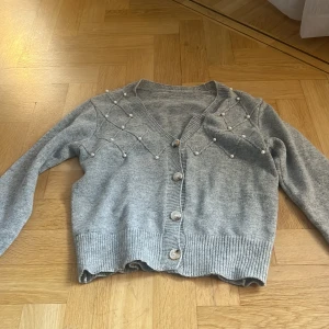 Grå kofta med pärldetaljer - Superfin grå kofta med v-ringning och snygga pärldetaljer på axlarna. Tröjan har ribbade muddar, knappar framtill och är stickad i mjukt material. Perfekt för dig som vill ha en söt och trendig look med det lilla extra.