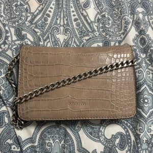 Beige axelväska i crocopräglat skinn - Snygg beige axelväska från Don Donna i crocopräglat skinnimitation med silverfärgad kedja. Väskan har ett foder med svartprickigt mönster och ett praktiskt innerfack med dragkedja. Perfekt storlek för det viktigaste och ger en cool vibe till din outfit.