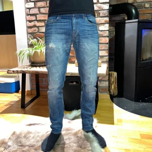 Blå jeans med slitningar från Tiger of Sweden - Säljer ett par blå jeans med slitningar på framsidan. De har en klassisk femficksdesign och en normal passform. Perfekta för en avslappnad stil.