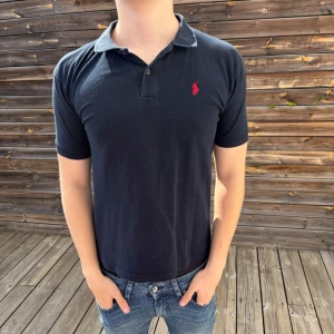Mörkblå piké från Ralph Lauren - Klassisk Mörkblå piké från Ralph Lauren med röd broderad logga på bröstet. Tröjan har krage, knappar vid halsen och korta ärmar. Tillverkad i mjuk bomull som känns skön mot huden. Perfekt för en clean och stilren look. Storlek S
