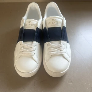 Valentino Garavani Open sneakers vit/blå - Snygga Valentino Garavani Open sneakers i vitt skinn med bred marinblå läderdetalj över mitten. Klassisk rund tå, vita snören och diskret logga i guld på plösen. Perfekta för dig som gillar stilrena och lyxiga sneakers med en modern twist.