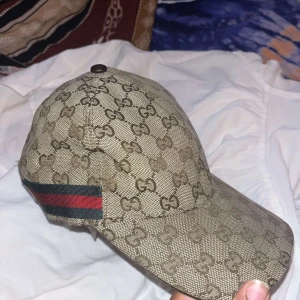Beige Gucci keps med GG-mönster - Säljer en beige keps från Gucci med klassiskt GG-monogram över hela kepsen och en ikonisk grön-röd rand på sidan. Kepsen är tillverkad i bomullscanvas och har böjd skärm. Perfekt för dig som vill sticka ut med en lyxig accessoar pris går att diskuteras.