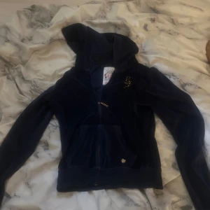 Mörkblå velourjacka från Lipsy - Mörkblå velourjacka med huva från Lipsy, dekorerad med guldiga nitar och stort LIPSY-tryck i hjärtform på ryggen. Jackan har dragkedja framtill, fickor och långa ärmar. Perfekt för dig som gillar en glam touch på din vardagsstil.