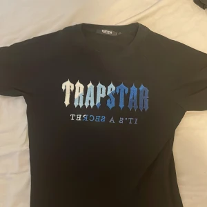 Svart Trapstar t-shirt med blått tryck - Svart t-shirt från Trapstar med ikoniskt tryck i vitt och blått på bröstet där det står 'TRAPSTAR IT'S A SECRET'. Klassisk passform och rund halsringning, perfekt för dig som gillar streetwear och vill sticka ut.
