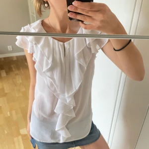 Vit blus med volangdetaljer - Supersöt vit blus med volanger längs fram och på axlarna. Blusen är ärmlös och har en lätt, luftig känsla som passar perfekt till sommaren. Snygg att styla med jeansshorts eller kjol för en fräsch look.