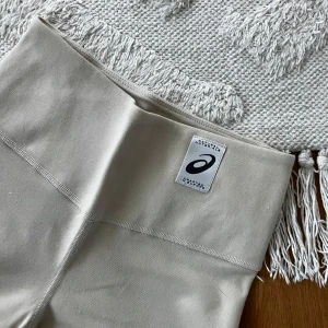 Beige leggings från Asics - Snygga beige tighta byxor med utsvängda ben från Asics med hög midja och stilren design. Tillverkade i mjukt och stretchigt material. Nypris 600kr