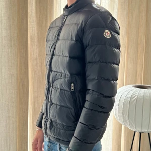Svart Moncler dunjacka herr - Säljer en svart dunjacka från Moncler med klassisk quiltad design och diskret logga på ärmen. Jackan har dragkedja framtill och två fickor med dragkedja. Perfekt för dig som vill ha en lätt och stilren jacka med premiumkänsla.