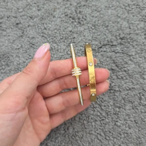 Två guldiga armband med stenar - Säljer två snygga armband i guldig metall. Det ena är smalt med glittrande stenar runt hela och flera ringar i mitten, det andra är bredare med infällda stenar och ingraverade symboler. Perfekta för att ge din outfit extra bling!