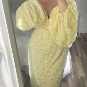 Ljusgul långklänning med puffärmar från adoore - Superfin ljusgul långklänning med små blommor, djup v-ringning och stora puffärmar. Klänningen har knappar framtill och en slits nertill. Perfekt för dig som vill sticka ut med en romantisk och somrig vibe.