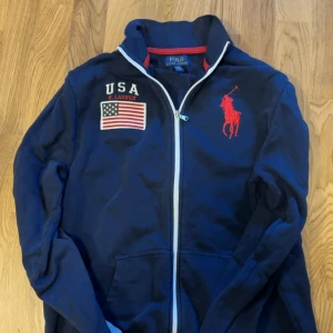 Blå ziptröja Polo Ralph Lauren  - Mörkblå ziptröja från Polo Ralph Lauren i storlek S . Jackan har broderad röd logga, USA-flagga och text på bröstet samt grå detaljer längs ärmarna. Tillverkad i mjuk bomull och har två fickor framtill. Perfekt för en sportig och avslappnad look.