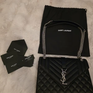 Svart quiltad axelväska Saint Laurent - Säljer en svart quiltad axelväska från Saint Laurent med ikoniskt YSL-spänne i silver. Väskan är i skinn med kedjerem och läderdetaljer, har innerfack med dragkedja och ytterfack. Kommer med dustbag och äkthetskort. Perfekt för dig som vill ha en lyxig och tidlös accessoar.