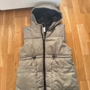 Beige dunväst med huva från Page One - Snygg beige dunväst från Page One Young med huva och svarta detaljer. Västen har dragkedja framtill, två stora fickor och justerbar snörning i midjan. Perfekt för lager-på-lager och chill höst- eller vinterstil.