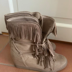 Beiga mockaboots med fransar - Snygga boots i beige mocka med massor av fransdetaljer runt skaftet och framtill. Skorna har rund tå och platt sula, perfekt för dig som vill sticka ut med en boho-inspirerad look. Lätta att matcha till jeans eller klänning. Som ni tyvärr ser på den sista bilden så har det gått upp lite på skorna 