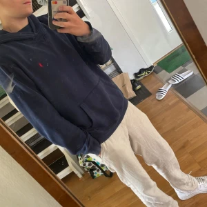 Mörkblå hoodie från Polo Ralph Lauren - Mörkblå hoodie från Polo Ralph Lauren med klassisk röd logga broderad på bröstet. Tröjan har huva, känguruficka och långa ärmar. Skön passform och tillverkad i mjuk bomull, perfekt för chill dagar.