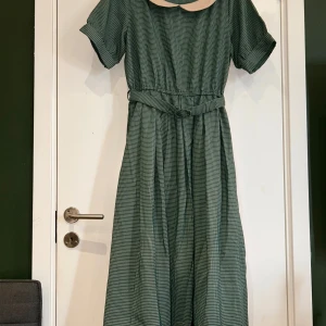 Grön rutig klänning med krage och bälte - Köpt i Japan, märke F.I.N.T.  Size: 36-38.  Supersöt långklänning i grönt och vitt rutigt tyg med korta ärmar, avskuren midja och matchande bälte. Klänningen har en ljus, rundad krage som ger en vintagevibe. Perfekt för dig som gillar retrostil och vill sticka ut lite extra.