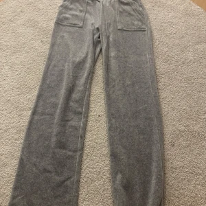 Grå mjukisbyxor Juicy Couture S - Grå mjukisbyxor från Juicy Couture i storlek S. Byxorna har bred resår i midjan med snörning, stora fickor fram och bak med knappdetaljer. Tillverkade i mjukt velourmaterial för en chill och trendig look.