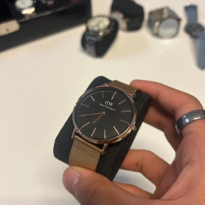 Svart och guldig klocka Daniel Wellington - Stilren klocka från Daniel Wellington med svart urtavla och guldig mesh-länk. Minimalistisk design med tunna guldiga visare och index. Perfekt accessoar för dig som gillar klassisk och modern stil. Priset är inte hugget i sten