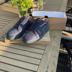 Mörkblå mocka sneakers från Louboutin - Exklusiva mörkblå sneakers från Christian Louboutin i lyxig mocka med klassisk röd sula och diskret logga på sidan. Skorna har rund tå, snörning och platt sula. Perfekta för dig som vill sticka ut med stilrena detaljer och premiumkänsla. Perfekt om du vill uppgradera din garderob för sommaren.