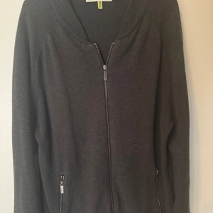 Mörkgrå kofta från Strömberg med dragkedja - Snygg grön/grå  kofta från Strömberg med hel dragkedja framtill och två fickor med dragkedja. Ärmarna har ribbade detaljer som ger en schysst look. Perfekt för lager-på-lager och funkar året runt. Materialet känns mjukt och skönt mot huden.