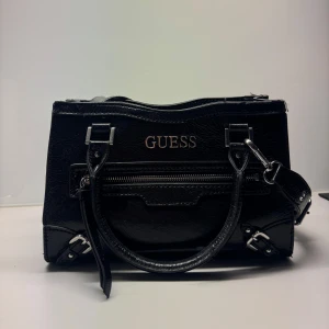 Svart handväska från Guess i skinn (guess traymore satchel) - Snygg svart handväska från Guess i glansigt skinn med silvriga detaljer. Väskan har både handtag och justerbar axelrem, dragkedja framtill och flera fack inuti. Perfekt storlek till vardags, och rymmer väldigt mycket. Väldigt bra skick, knappt använt.