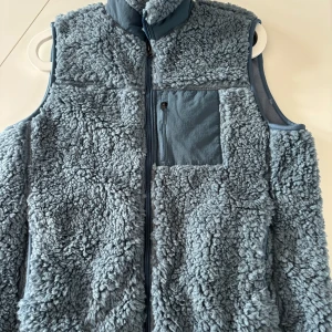 Blå fluffig fleeceväst med dragkedja - Mysig blå fleeceväst med fluffig utsida och mjuk insida. Västen har hög krage, dragkedja framtill och en praktisk bröstficka med dragkedja. Perfekt lager-på-lager-plagg för kyliga dagar och riktigt skön att ha på sig. Stl 160. Helt ny. 