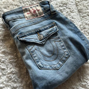 True Religion - Säljer ett par ljusblå skinny jeans från True Religion med slitna detaljer, dekorativa knappar och ikoniska bakfickor. Storlek: 28, stretchigt material. 