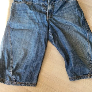 Blå jeansshorts med bakficka - Snygga blå jeansshorts med klassisk femficksdesign och en stor broderad C-detalj på bakfickan. Shortsens denim är i en mellanblå tvätt med slitningar och kontrastsömmar. Perfekta för en chill sommarlook.