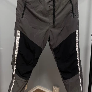 Grå och svarta byxor från American Eagle - Snygga grå och svarta byxor från American Eagle med vita logoremsor längs sidorna. Byxorna har dragkedjefickor, snörning i midjan och är gjorda i ett lätt syntetmaterial. Perfekta för en sportig och avslappnad stil.