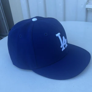 Blå LA-keps New Era 59FIFTY 7 1/4 - Snygg blå keps från New Era 59FIFTY med LA-logga broderad i vitt framtill. Klassisk rak skärm och vit knapp på toppen. Tillverkad i polyester, perfekt för dig som gillar sportig stil och vill sticka ut med en ikonisk look.