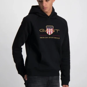 Svart hoodie från GANT med tryck - Svart hoodie från GANT med stort broderat logotyp-tryck och sköld på bröstet. Klassisk känguruficka framtill, huva och långa ärmar. Mjuk insida och sportig vibe, perfekt för chill dagar. Snygga detaljer i guld och rött på trycket. Säljer då min lillebror fick den i julklapp men han har aldrig använt den