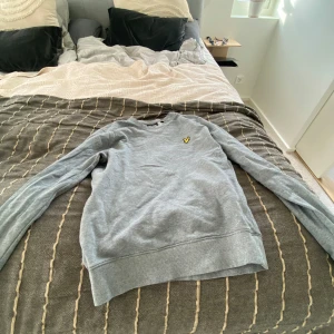 Grå sweatshirt från Lyle & Scott - Snygg grå sweatshirt från Lyle & Scott med klassisk rund hals och broderad gul logga på bröstet. Tröjan är i mjuk bomull och har långa ärmar samt ribbade muddar vid ärmslut och nederkant. Perfekt för en chill och stilren look.