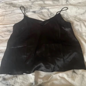 Svart linnetopp i satin från Lager 157 - Snygg svart linnetopp från Lager 157 i glansigt satinliknande material. Topp med tunna axelband och v-ringad halsringning, perfekt att styla med jeans eller kjol. Enkel och stilren design som ger en lyxig känsla.