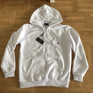 Vit hoodie från Polo Ralph Lauren - Säljer en klassisk vit hoodie från Polo Ralph Lauren med dragkedja och huva. Tröjan har två fickor framtill och den ikoniska broderade loggan på bröstet. Perfekt för en stilren och avslappnad look. Tröjan är i storlek S men Passar även M. Tröjan är bara testad. Priset är ej hugget i sten!