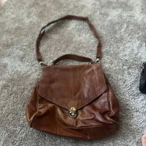 Brun axelväska i skinn från Vera - Snygg brun axelväska i äkta skinn från Vera. Väskan har ett klassiskt lock med metallspänne, justerbar axelrem och praktiskt handtag. Invändigt finns flera fack och en dragkedja för småsaker. Perfekt för dig som vill ha både stil och funktion.