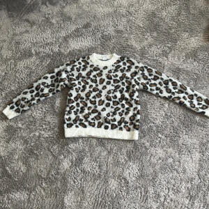 Leopardmönstrad stickad tröja - Säljer en fluffig stickad tröja med leopardmönster i svart och brunt på beige botten. Tröjan har rund halsringning, långa ärmar och ribbade muddar. Perfekt för dig som vill sticka ut med en trendig och djurinspirerad look.