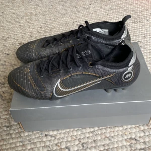 Nike Vapor 14 Elite AG fotbollsskor - Snygga Nike Vapor 14 Elite AG fotbollsskor i svart med metallic silverdetaljer och guldiga linjer. Skorna har snörning, låg profil och är tillverkade i syntetmaterial för bästa grepp och känsla på planen. Perfekta för dig som vill ha stil och prestanda på fotbollsplanen.