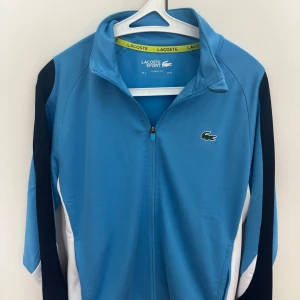 Blå vindjacka från Lacoste Sport - Snygg blå kofta från Lacoste Sport med dragkedja framtill och klassisk krage. Jackan har vita och mörkblå detaljer längs sidorna och ärmarna samt den ikoniska krokodilloggan på bröstet. Perfekt för en sportig och stilren look.