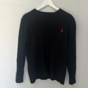 Svart kabelstickad tröja Polo Ralph Lauren - Svart kabelstickad tröja från Polo Ralph Lauren med klassisk röd broderad logga på bröstet. Tröjan har rund halsringning och långa ärmar, perfekt för lager-på-lager. Mjuk och skön känsla, passar till allt och ger en clean look.