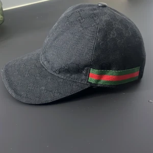 Svart keps från Gucci med GG-mönster - Snygg svart keps från Gucci med diskret GG-monogram över hela tyget. Klassisk böjd skärm och justerbar rem bak. Sidorna har den ikoniska gröna och röda randen. Tillverkad i bomullstyg med exklusiv känsla.