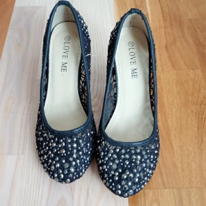 Svarta pumps med glittriga stenar - Snygga svarta pumps från Love Me med spetsdetaljer och massor av glittriga stenar över hela skon. Skorna har rund tå och hög klack, perfekt för dig som vill sticka ut. Ovandelen är i spets och syntetmaterial för en elegant look.