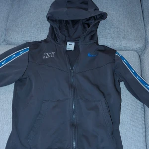 Svart Nike kofta med blå detaljer - Svart Nike kofta  med huva och dragkedja. Ärmarna har blå och vita Nike-loggor längs sidorna. Jackan har två fickor framtill och Dri-FIT-tyg som andas. Perfekt för dig som gillar sportig stil och vill ha något bekvämt och snyggt.