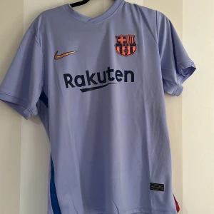 FC Barcelona bortatröja Nike ljusblå - FC Barcelona bortatröja från Nike i ljusblått med klubbmärke på bröstet och Rakuten-tryck framtill. Tröjan har korta ärmar, rund hals och är tillverkad i ventilerande polyester. Snygg detalj med blå rand på sidan och UNICEF-tryck på ryggen. Kan diskutera pris
