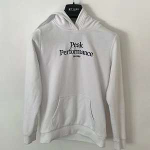 Vit hoodie från Peak Performance - Säljer en stilren vit hoodie från Peak Performance med svart broderad logga på bröstet. Tröjan har huva, känguruficka och långa ärmar. Perfekt för dig som gillar en clean och sportig look.