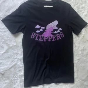Svart Steppers t-shirt med lila tryck - Svart t-shirt från Steppers med ett stort lila tryck framtill som visar en kopp som häller ut vätska bland moln och texten 'STEPPERS'. Klassisk passform och rund hals. Perfekt för dig som gillar streetwear och vill sticka ut med cool grafik.