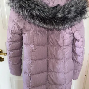 Ljuslila dunjacka från The North Face - Säljer en lång, ljuslila dunjacka från The North Face med snyggt mönstrat tyg och stor huva med grå fuskpäls. Jackan har dragkedja och knappar framtill, quiltad design och är perfekt för kalla dagar. Riktigt mysig och stilren vinterjacka.
