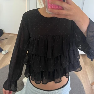 Svart volangblus från Gina Tricot - Supersöt svart blus från Gina Tricot med små prickar i tyget och flera lager volanger framtill. Blusen är genomskinlig med lång ärm och knapp i nacken. Perfekt för dig som gillar en romantisk och trendig look. som inte säljs i butiken längre. Säljer därför att den är för kort.